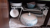 Photo showing stacked turquoise blue dinnerware plates, cups on rack, and white covered casserole dish with decorative raised relief and turquoise detailing