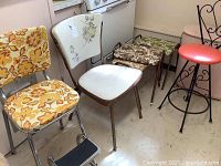 Front view of four vintage kitchen chairs with vinyl upholstery including floral patterns and a metal bar stool with red vinyl seat, small stools with floral cushions.