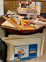 Wooden T-squares, sketch pads, drawing boards, watercolor block, variety of art supplies arranged on a round table.