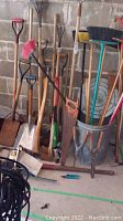 Wide view showing assortment of rakes, shovels, hoes, brooms leaning against cinderblock wall with metal bucket holding hand tools on floor