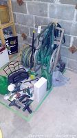 Wide view showing garden hoses on reels, watering cans, and sprinklers grouped on floor against wall.