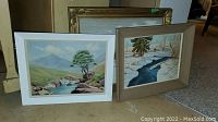 Three oil paintings propped against a wall, two frames visible (white and light brown), scenes are natural landscapes with water and trees, varying sizes.