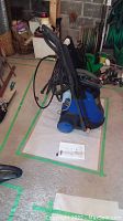 Blue and black Simonize pressure washer with hoses, power cord, and handle in garage setting.
