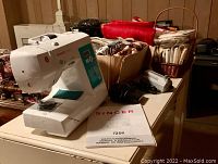 White Singer sewing machine model 7258 on a table with instruction manual beside it. Various packages and boxes in the background containing sewing supplies and fabrics.