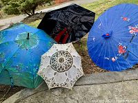 Three umbrellas displayed open on ground: blue painted Monet-style, black London-themed with red details, white crochet parasol.