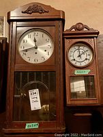 Two vintage wooden pendulum clocks side by side showing front overall view. Larger clock with silver dial and smaller clock with Roman numeral dial.