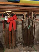 Two wooden sleds side by side leaning against a wooden board, one decorated with a large red bow and showing rust on metal parts.