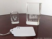 Both etched crystal paperweights displayed on a dark surface, showing the smaller eagle design and larger New York City skyline design.