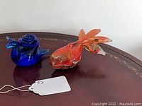 Both glass animal figurines placed on a wooden surface with a white tag visible. The colorful angel fish is on the right and the cobalt blue snail on the left.