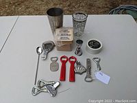 Overview of barware assortment showing cocktail shaker, bottle openers, mixing spoon, strainer, glass, jigger, cocktail napkins, and container.
