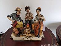 Three men in porcelain sculpture playing music instruments and drinking wine with detailed base, viewed front left angle.