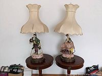 Pair of porcelain Capodimonte style figural table lamps with fabric shades placed on wooden tables.