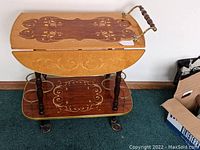 Front view of the bar cart showing the wooden drop leaf top with intricate floral inlay and brass edging, four turned legs, bottom shelf with matching inlay, and caster wheels.