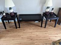 Photo showing the pair of black square contemporary end tables alongside a black rectangular coffee table and decorative ceramic lamps. Wood grain flooring visible beneath.