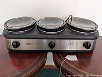 Front view of the triple buffet slow cooker showing three ceramic pots with glass lids and three control knobs.