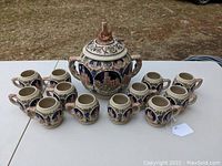 Full set view of the large stoneware punch bowl with lid surrounded by 12 matching mugs arranged on a table outdoors.