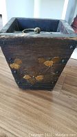 Front side showing carved and painted leaf and flower motif with metal ring handle on top.