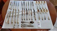 Full set of cutlery laid out on a table showing knives, spoons, butter cutters, small spoons, and scissors arranged in groups.