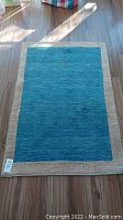 Full view of blue area rug with beige border lying flat on wood floor, showing overall shape and colors.