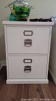 Front view of white filing cabinet with two drawers and metal handles, holding decorative items on top.