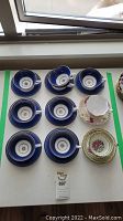 Eight Mintons Washington teacups and seven matching saucers, one floral Queen Anne teacup and saucer, one white and gold Foley teacup and saucer all laid out on a table.