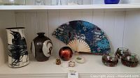 Wide shot of entire lot showing signed crow vase, secondary pottery vase, Japanese fan, red decorative ball, painted stones, resin monk, and glass bowls with floral decor