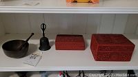 Metal singing bowl with wooden striker, metal bell, two red lacquer carved boxes on white shelf