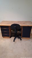 Front view of pine wood desk with black drawers and cabinet alongside a black rolling office chair.