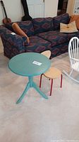Shows the round teal painted pedestal table with accompanying small wooden chair in front of a patterned sofa