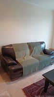 Chocolate brown leather sofa covered partially with a green throw, showing wooden feet and armrest detail.