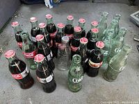 Overview of approximately 20 assorted Coca-Cola glass bottles, including early 1900s embossed clear bottles and green bottles with caps.