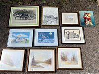 Full view of all nine framed artworks arranged outdoors on gravel showing various subject matter and frame styles.