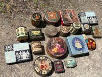Main photo showing assorted tins grouped on ground outside with a variety of shapes and decorative designs visible including flowers, children, classical figures, and rustic nature scenes.