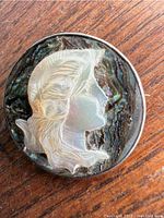 Front view of round carved cameo brooch with delicate profile carved from mother of pearl on abalone shell backing showing iridescent blues, greens, and purples. Mounted in sterling silver.