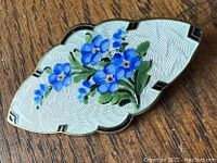 Top and front view of curved shield-shaped enamel brooch with detailed blue flower cluster on white guilloché background resting on wood surface.