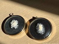 Two oval floral cameos in gold-tone round frames on black velvet background, displayed side-by-side in natural light showing detail and size