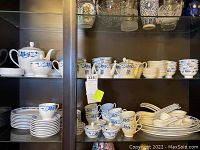 Glass cabinet showing blue and white china dinnerware including stacked plates, cups, bowls, serving dishes and covered containers.