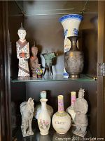 Display of Asian figurines and decorative vases on glass shelves, showing various sizes and materials including wood, bronze, porcelain, and cloisonné.