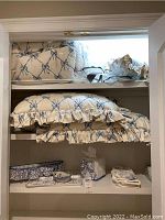 Shelf with quilted comforter, pillow shams, pillows, folded linens, and tissue box holder all decorated in blue ribbon bow pattern with ruffled edges.