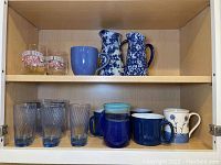 Full view of cabinet shelf showing 6 mugs including handmade signed, 2 pitchers with blue floral designs, 7 blue juice glasses and 2 stemless wine glasses.