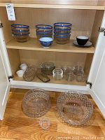 Full view of all items in wooden cabinet, including blue rim glass bowls, Noritake cup, ramekins, crystal bowls and various glass serving pieces.