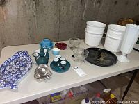 Full overview of the lot on two tables, showing the arrangement of all ceramic, glass, metal, and pottery items including the fish shaped plate, white vases, green pitchers and cups, metal bowls, and glass bowls.