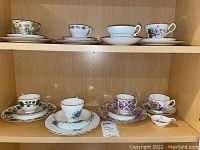 Overall view of eight bone china teacups and saucers with six matching dessert plates and one tea leaf saucer on wooden shelves, showing variety of floral patterns and gold trims.