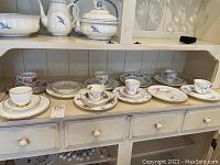 Wide view of all teacups, saucers, and plates arranged on a cream-colored shelf and tabletop, showing pattern variety and item counts.