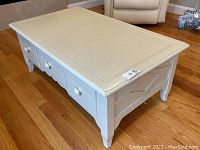 Top and front view of an off-white painted wood coffee table with three drawers and round knobs, showing grooved panel design on top and some paint wear.