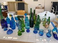 Full view of assortment of green and blue glass items including bottles, bowls, and figures arranged on a tiled white surface