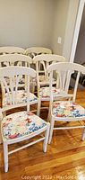 Six painted wood dining chairs arranged on a wood floor showing the floral patterned upholstered seats and white painted frames.