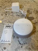 Top view of Amazon Echo Dot with four control buttons and white charging cord with adapter coiled beside it on a marble surface.