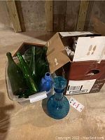 Photo showing a collection of various green and blue glass bottles and decanters on a floor, some in a plastic bin and others standing beside it.