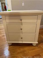 Front view of the white nightstand showing three drawers with round metal knob handles and bun feet on hardwood floor.